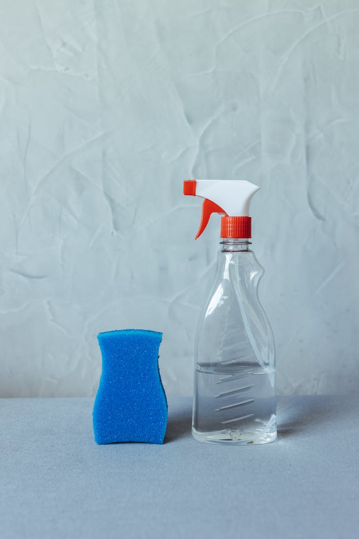 A clear spray bottle and blue sponge on a textured surface for cleaning tasks.