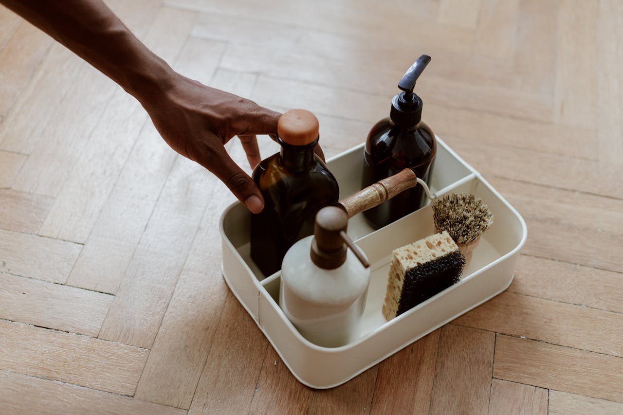A hand reaching for a cleaning set with bottles and brushes on a wooden floor.